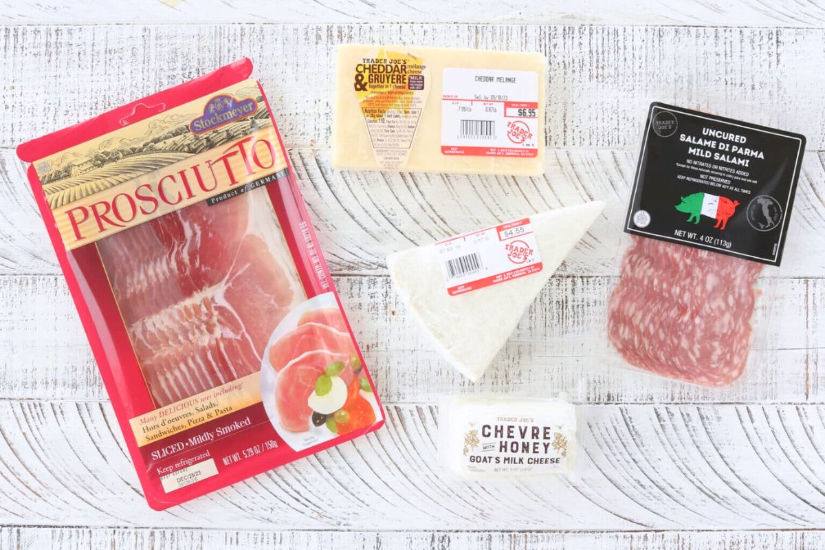 Ingredients for an easy charcuterie board - Prosciutto, Cheddar, Brie, Goat Cheese and Salami on a white board.