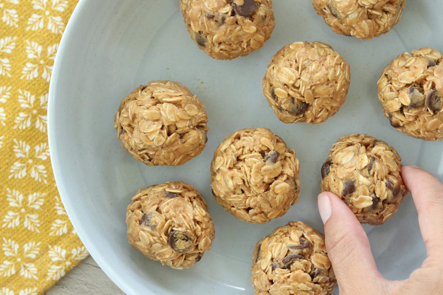 Hand reaching for a no-bake peanut butter oatmeal energy ball with chocolate chips from a plate of energy balls.