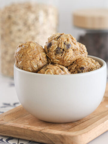 No bake peanut butter power balls with rolled oats and chocolate chips stacked in a white bowl on a wood board, with jars of oats and chocolate chips behind.