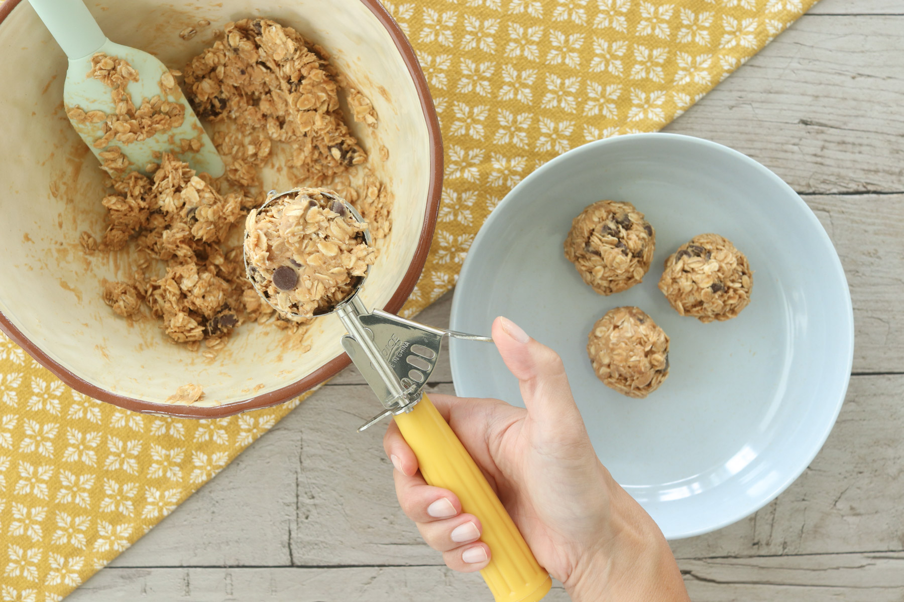 Hand using a cookie scoop to portion no-bake peanut butter power ball mixture, with rolled balls on a plate beside the mixing bowl.