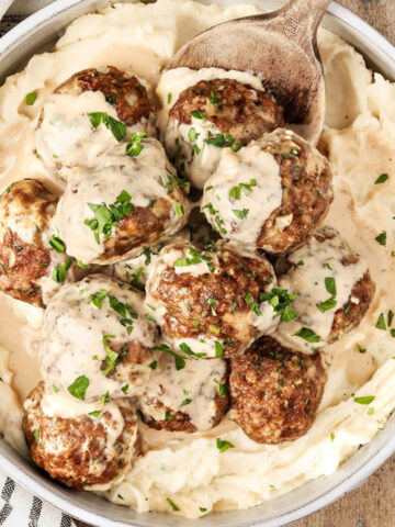 Overhead view of homemade Swedish meatballs covered in creamy gravy, served over mashed potatoes and garnished with fresh parsley in a white bowl on a rustic wooden table.