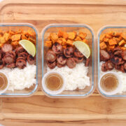 Three glass meal prep containers filled with white rice, roasted sweet potatoes, sliced chicken sausage, lime wedges, and honey lime dressing.