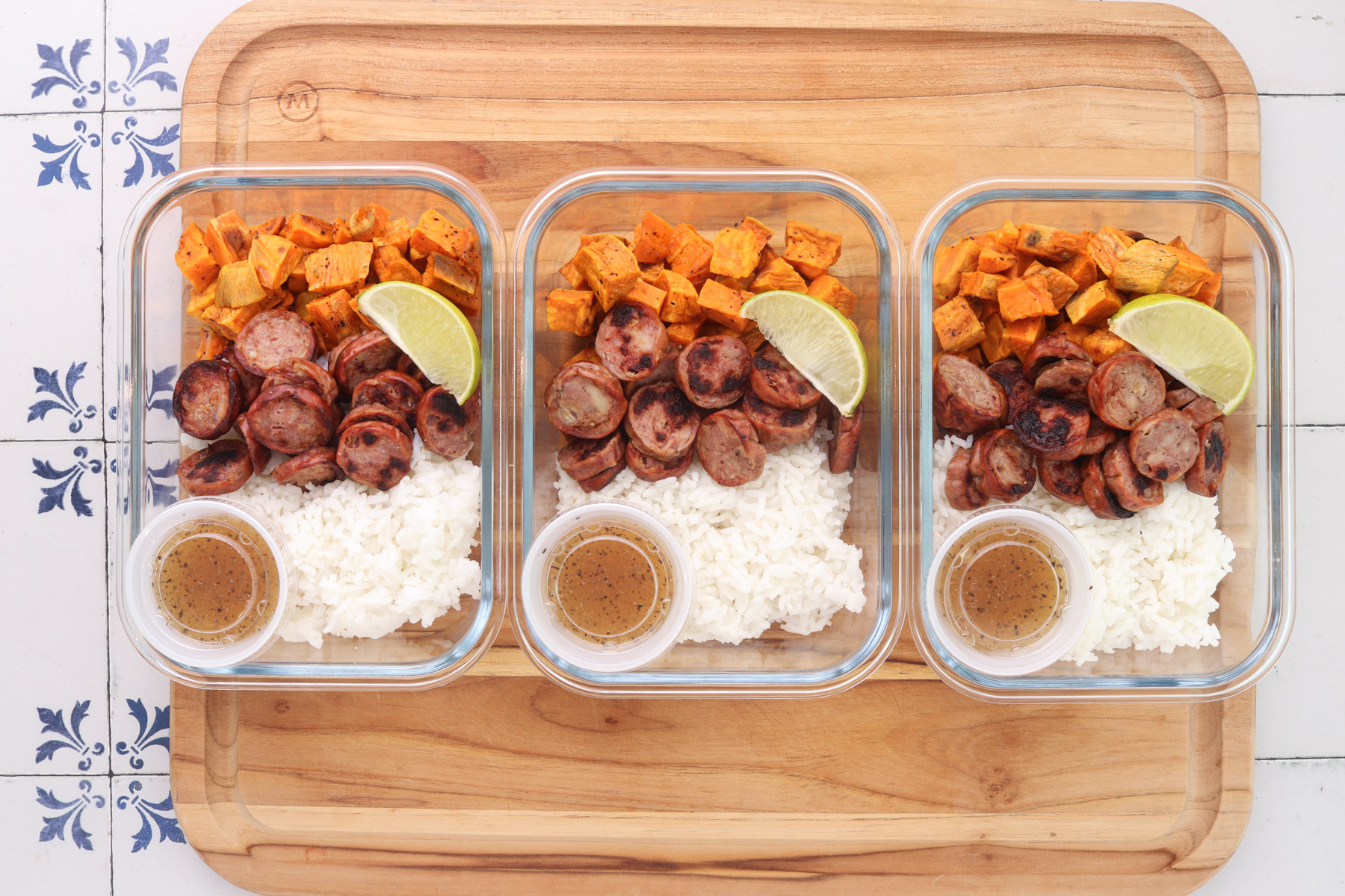 Three glass meal prep containers filled with white rice, roasted sweet potatoes, sliced chicken sausage, lime wedges, and honey lime dressing.
