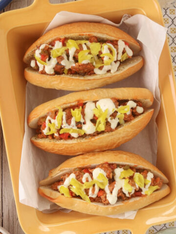 Three Italian sausage sandwiches stuffed with sausage, peppers, onions, and marinara, topped with creamy garlic-Parmesan sauce and pepperoncini on toasted rolls.