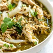 Close-up of a white bowl filled with shredded salsa verde chicken, garnished with fresh cilantro and sliced onions, with a light green broth visible around the chicken.