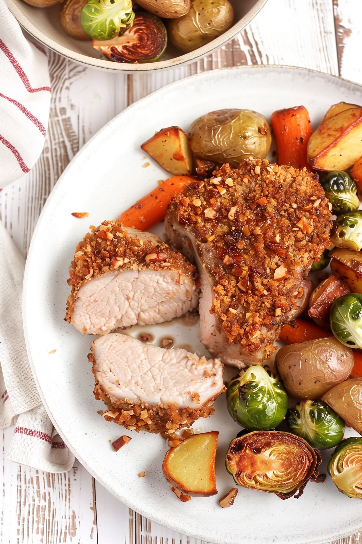 A white plate with pecan-crusted boneless pork chops, one sliced to show the juicy inside, served with roasted carrots, baby potatoes, and halved Brussels sprouts. The vegetables are golden and caramelized, and the pork chop has a crunchy pecan breadcrumb coating.