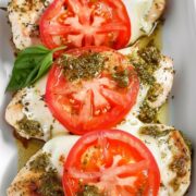 Overhead view of baked pesto chicken breasts topped with melted mozzarella, fresh tomato slices, and basil, arranged in a white ceramic baking dish.