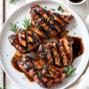 Overhead image of grilled chicken glazed with balsamic vinegar chicken marinade, served on a white plate with rosemary sprigs, a small bowl of marinade, and a rustic whitewashed wood background.