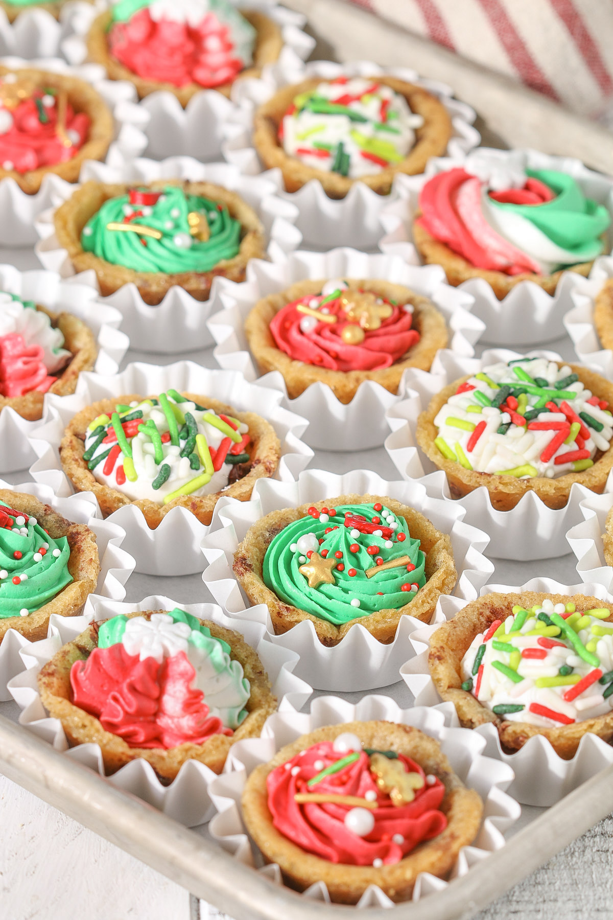 Overhead view of mini chocolate chip cookie cups baked in a mini muffin pan and filled with colorful buttercream frosting and Christmas sprinkles, arranged in white paper cups.