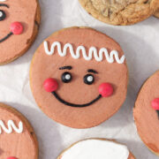 Close-up of decorated gingerbread-face sugar cookies with smooth brown frosting, pink cheeks, black piped smiles, and white icing details, arranged on parchment paper alongside plain chocolate chip cookies.