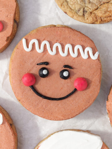 Close-up of decorated gingerbread-face sugar cookies with smooth brown frosting, pink cheeks, black piped smiles, and white icing details, arranged on parchment paper alongside plain chocolate chip cookies.