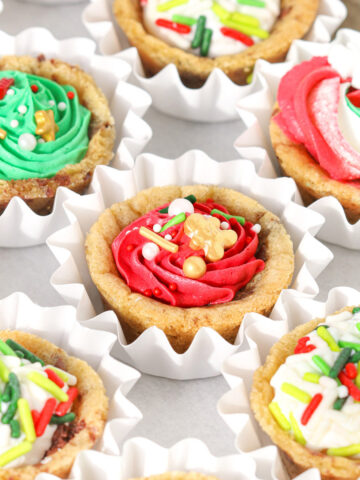 Overhead view of mini chocolate chip cookie cups baked in a mini muffin pan and filled with colorful buttercream frosting and Christmas sprinkles, arranged in white paper cups.