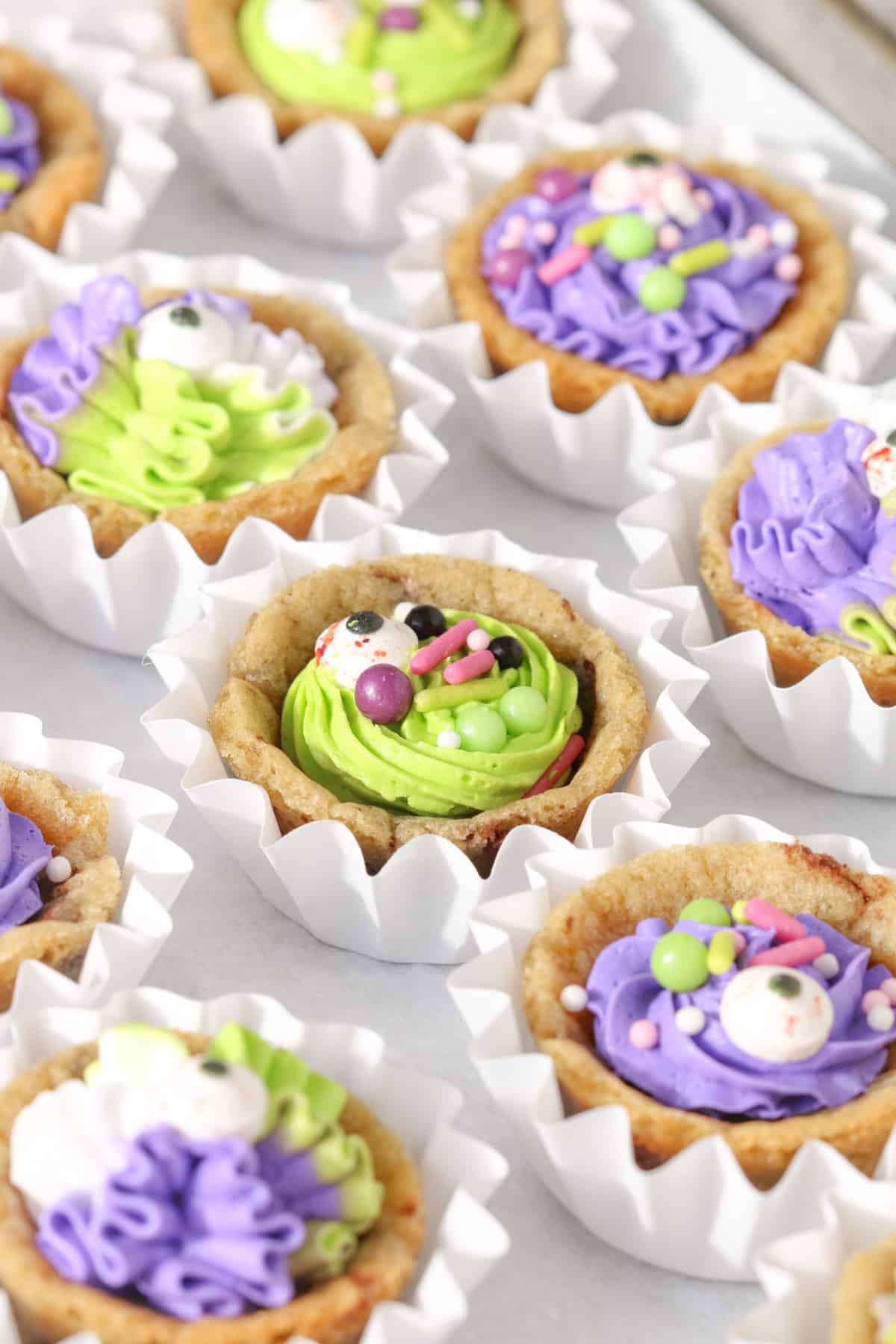 Mini chocolate chip cookie cups filled with bright green and purple buttercream and decorated with Halloween sprinkles and candy eyeballs, arranged in white ruffled paper liners.