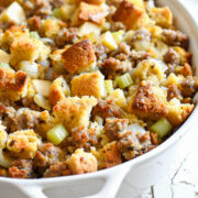Close-up of homemade sausage and apple stuffing in a white baking dish. The stuffing features golden cornbread cubes, diced celery, onions, and browned mild Italian sausage, baked until crisp on top and tender inside.