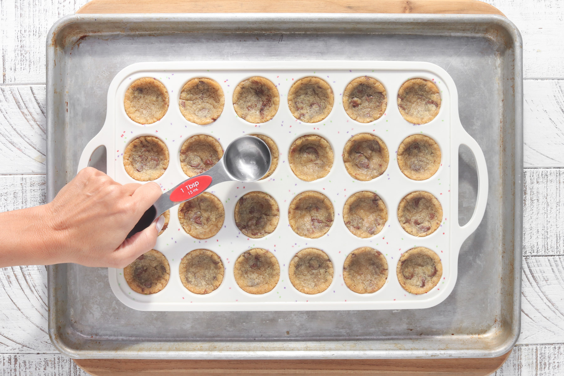 Hand pressing the center of baked cookie cups with a tablespoon in a silicone mini muffin pan to form cookie cup shapes.