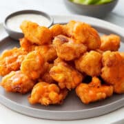 Buffalo cauliflower bites on a gray plate, coated in a light orange buffalo sauce with a soft baked batter, served with celery sticks and a small bowl of ranch dressing.