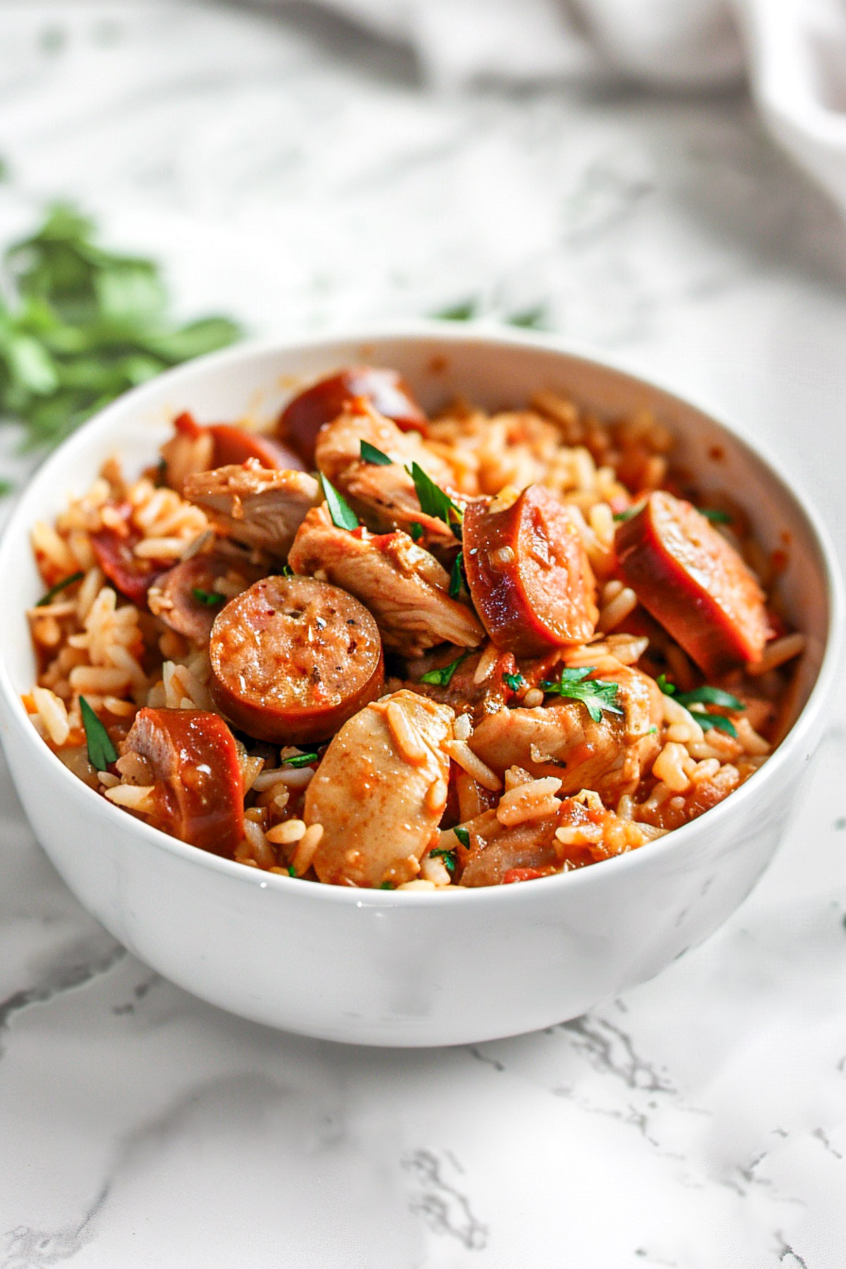 Bowl of chicken, sausage, and shrimp jambalaya with rice, garnished with chopped parsley on a light marble surface.