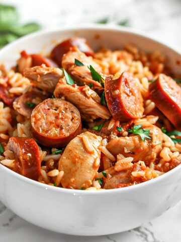 Bowl of chicken, sausage, and shrimp jambalaya with rice, garnished with chopped parsley on a light marble surface.