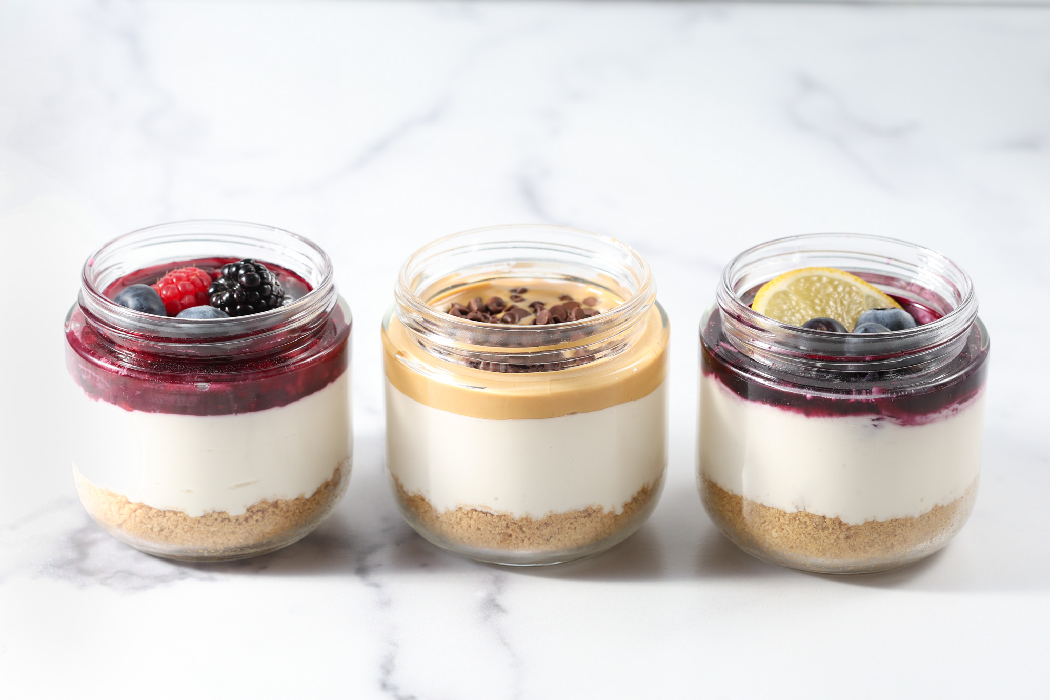 Three small glass jars filled with high-protein cheesecake, layered with graham cracker crumbs on the bottom and creamy cheesecake filling, topped with berry compote and fresh berries, peanut butter with chocolate chips, and blueberry lemon topping on a white marble surface.
