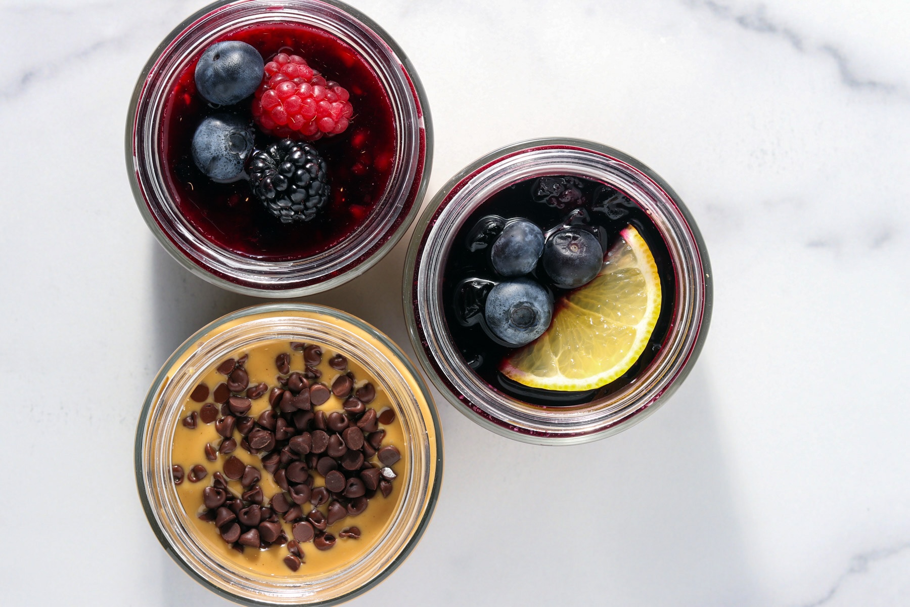 Overhead view of three high protein cheesecake jars with different toppings, including mixed berry compote with fresh berries, blueberry lemon topping with lemon slice, and peanut butter cheesecake topped with chocolate chips, arranged on a light marble surface.
