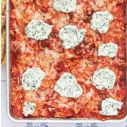 Sheet pan lasagna in an aluminum baking sheet, topped with dollops of herbed ricotta over melted mozzarella and red meat sauce, with a mustard yellow napkin at one corner.