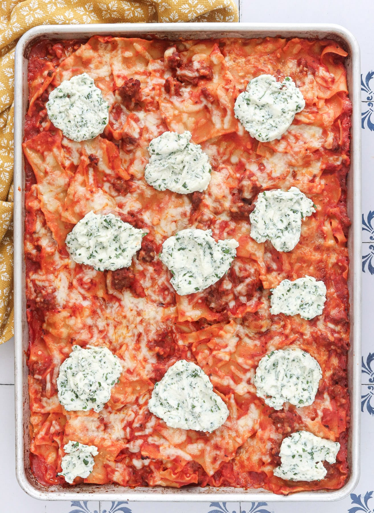 Sheet pan lasagna in an aluminum baking sheet, topped with dollops of herbed ricotta over melted mozzarella and red meat sauce, with a mustard yellow napkin at one corner.