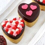 Mini heart-shaped cakes coated in smooth dark chocolate, decorated with red, pink, and white piped frosting, and topped with pink conversation-heart candies on a light wood surface.
