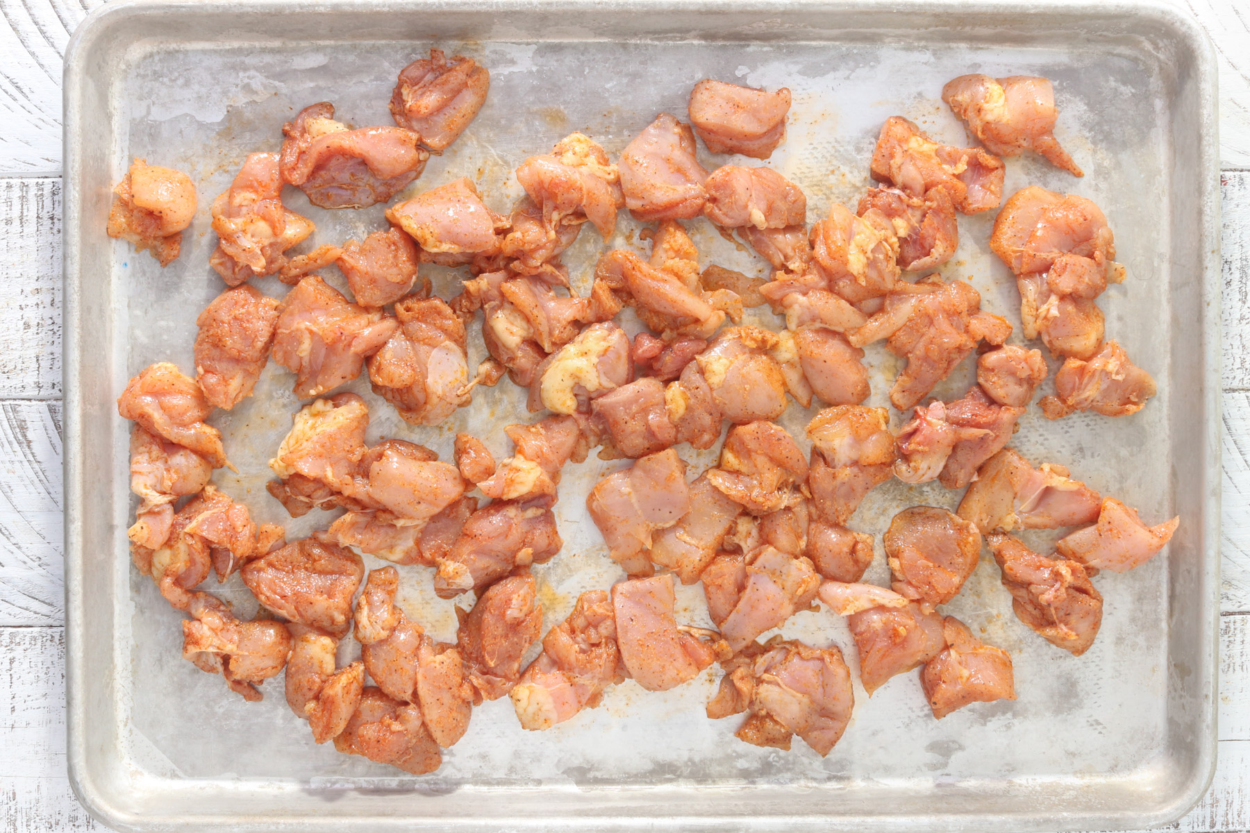 Raw diced chicken thighs tossed with oil and seasonings, spread in a single layer on a rimmed sheet pan before roasting.