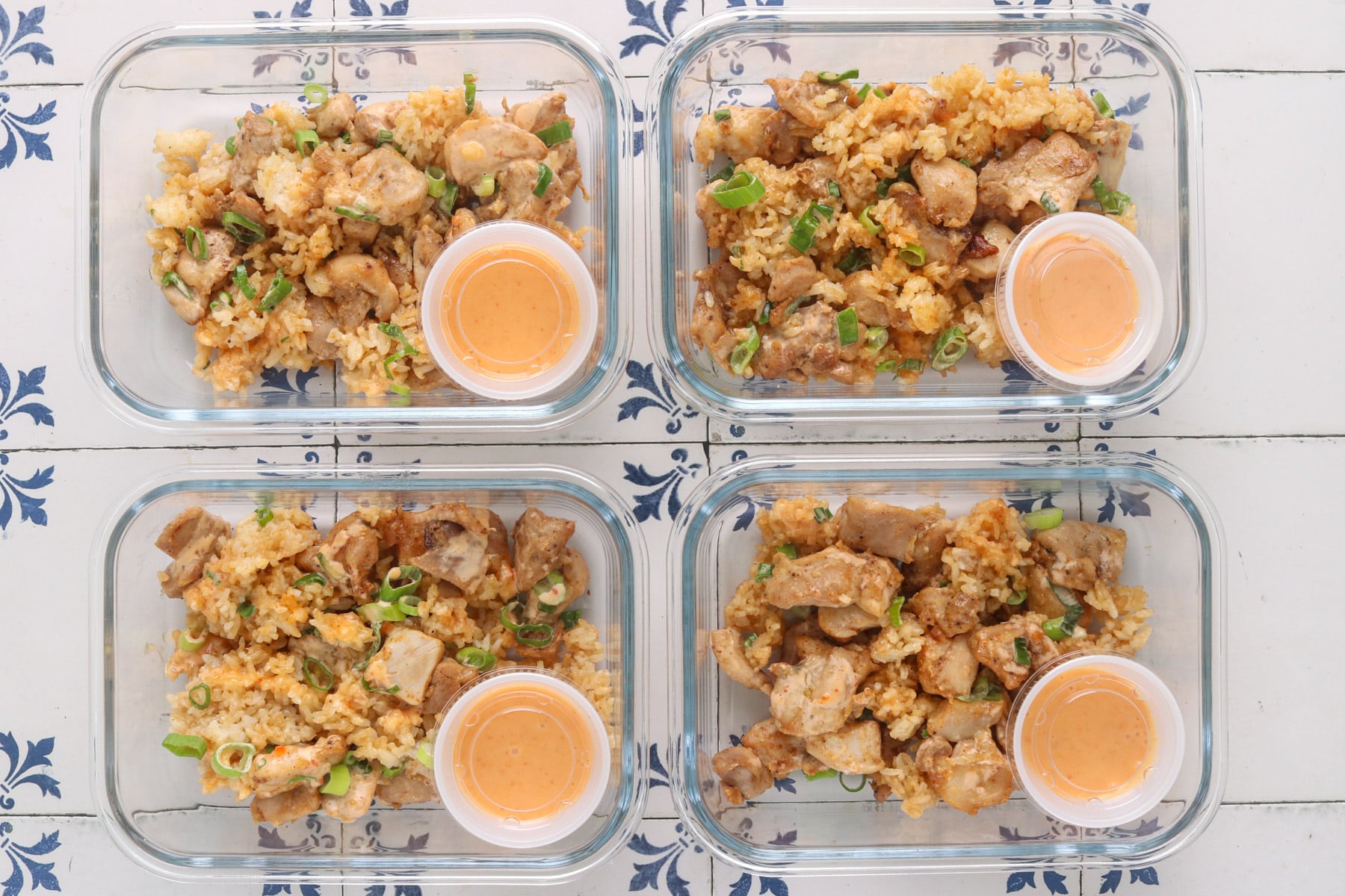 Bang bang chicken and crispy rice portioned into four glass meal prep containers with sliced green onions and bang bang sauce on the side.
