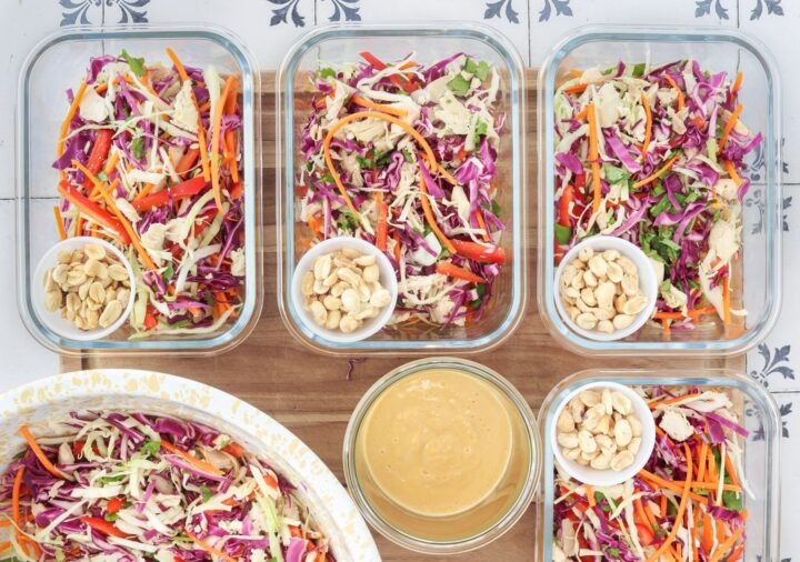Overhead photo of four glass meal-prep containers filled with shredded chicken, red and green cabbage, carrots, bell peppers, and cilantro, each with a small cup of peanuts. A bowl of additional salad and a jar of creamy peanut dressing sit on a wooden cutting board.