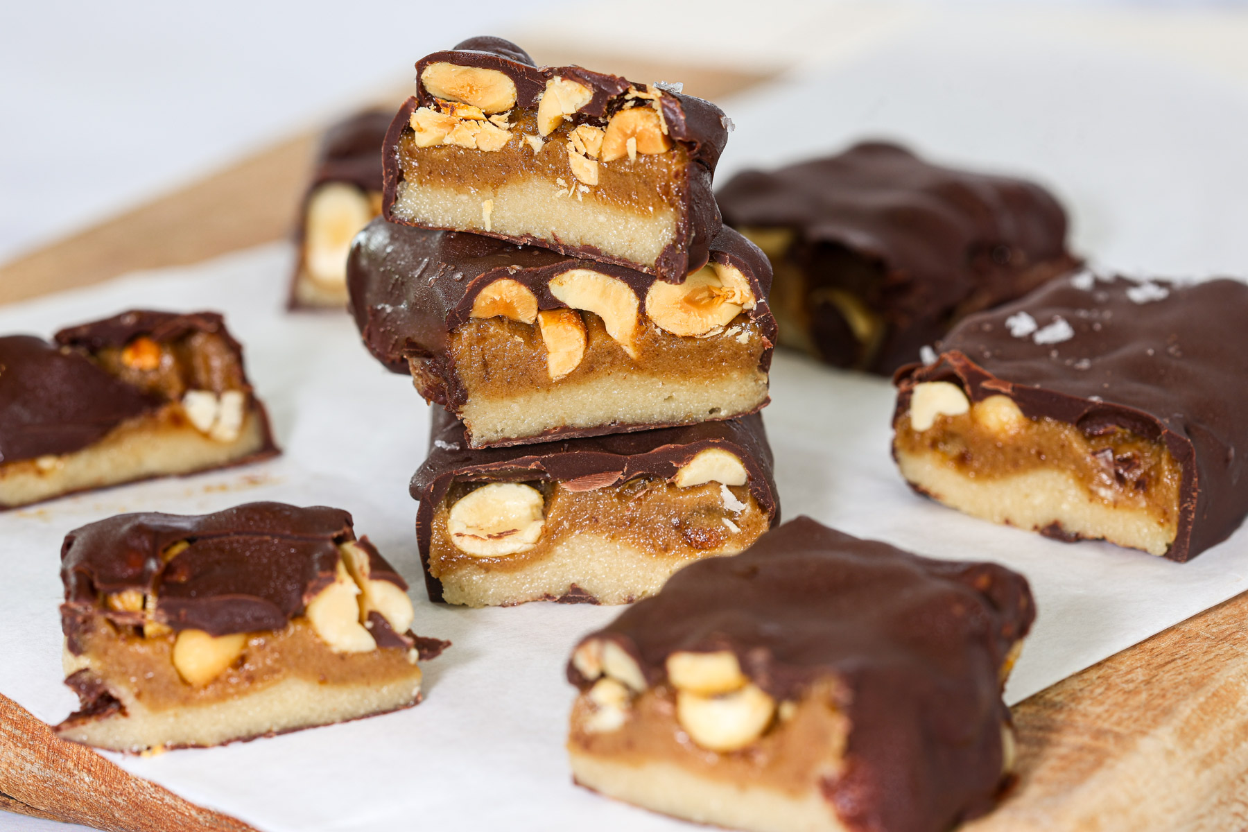 Stack of three healthy snickers bars cut to show layers of almond flour base, date caramel, and roasted peanuts under a dark chocolate coating.