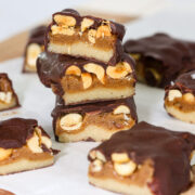 Stack of three healthy snickers bars cut to show layers of almond flour base, date caramel, and roasted peanuts under a dark chocolate coating.