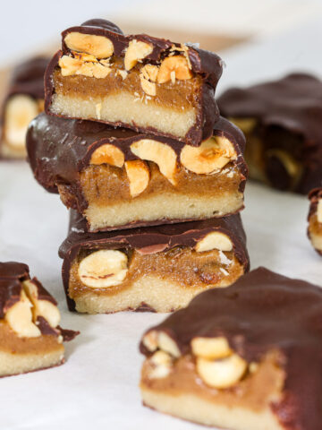 Stack of three healthy snickers bars cut to show layers of almond flour base, date caramel, and roasted peanuts under a dark chocolate coating.
