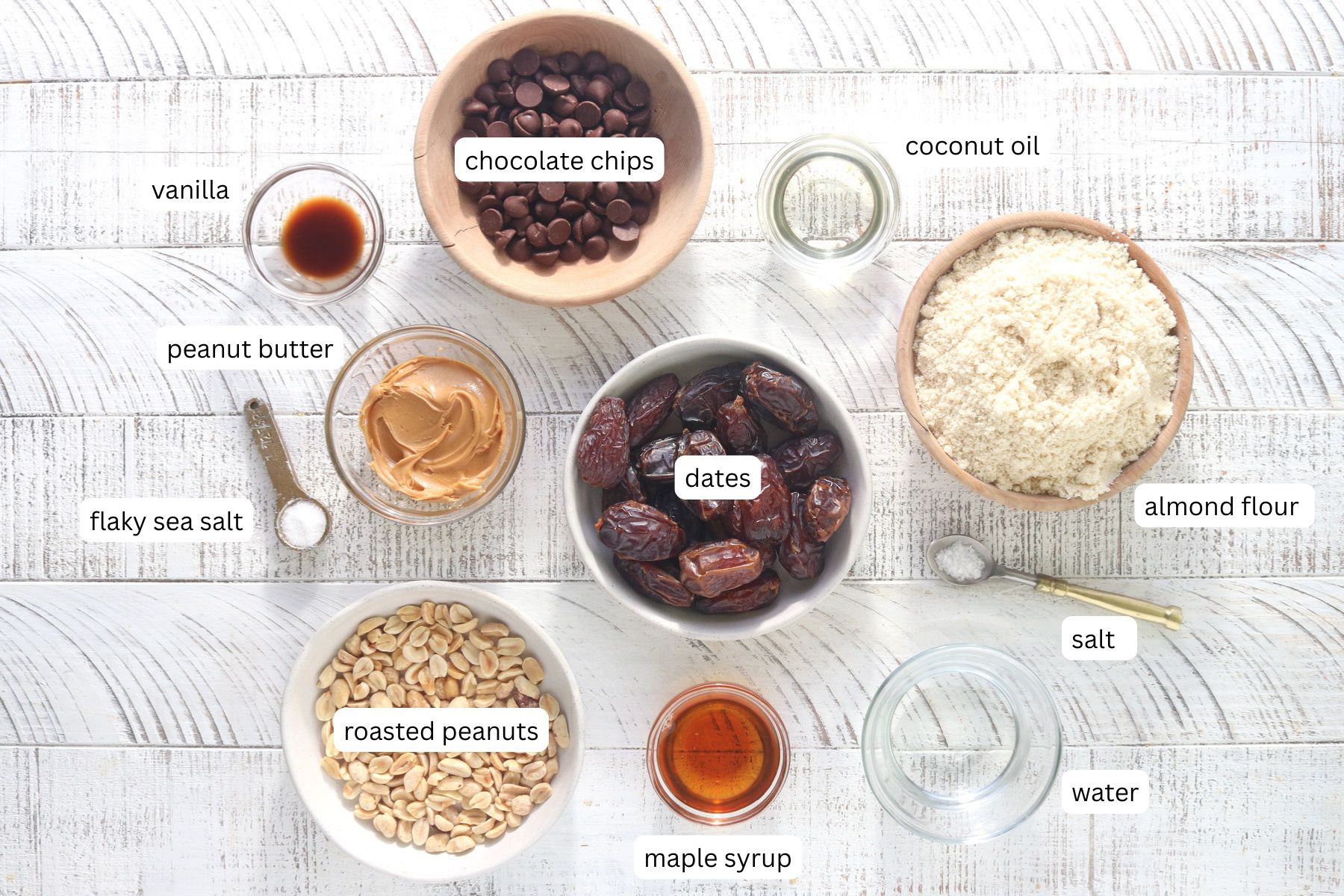 Ingredients for healthy snickers bars laid out and labeled, including almond flour, Medjool dates, peanut butter, dark chocolate chips, roasted peanuts, maple syrup, coconut oil, vanilla, salt, and water.