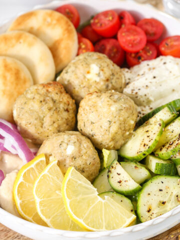 Mediterranean chicken meatball bowl with cucumber salad, cherry tomatoes, hummus, pita bread, red onion, and lemon wedges.
