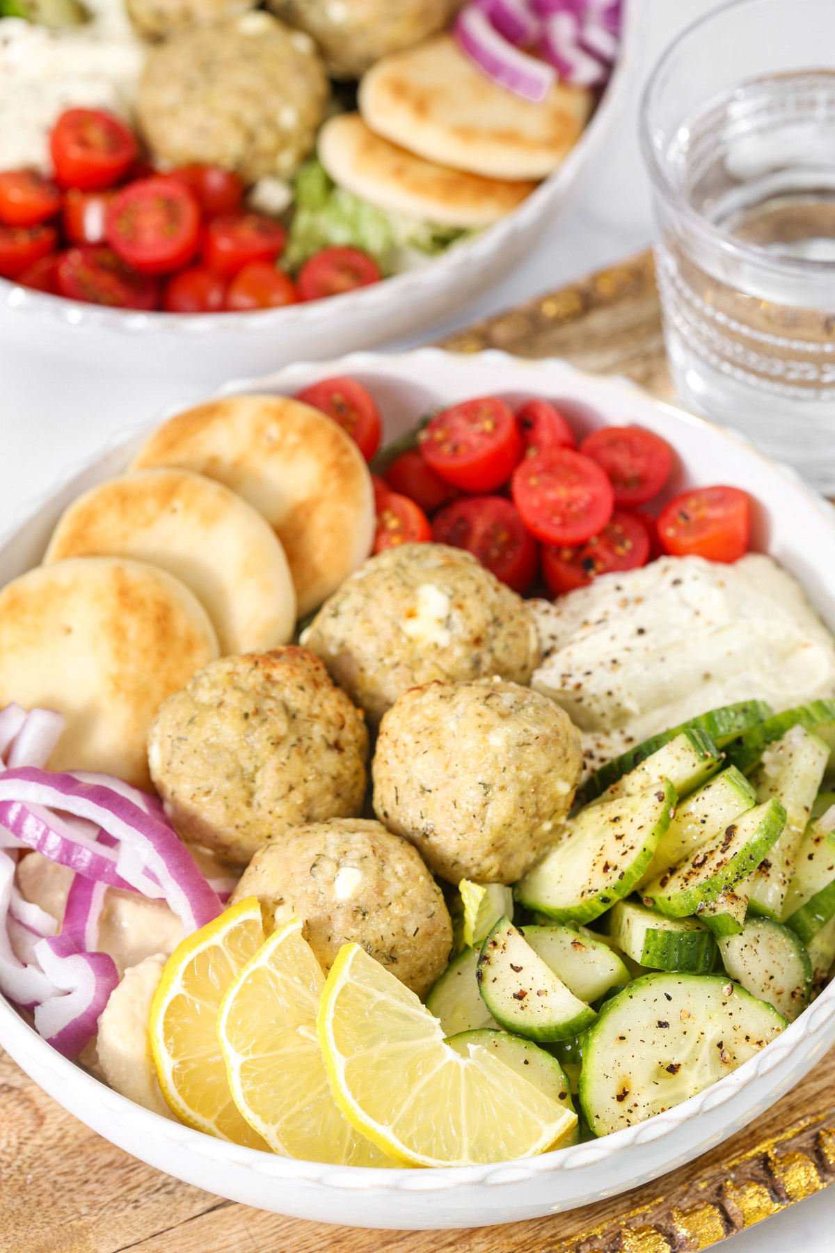 Mediterranean chicken meatball bowl with cucumber salad, cherry tomatoes, hummus, pita bread, red onion, and lemon wedges.