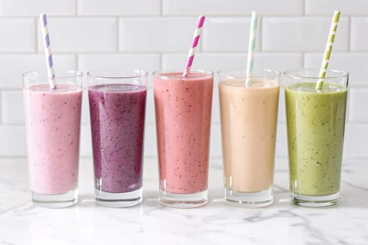 Five colorful smoothies in tall glasses with striped straws lined up on a marble counter.