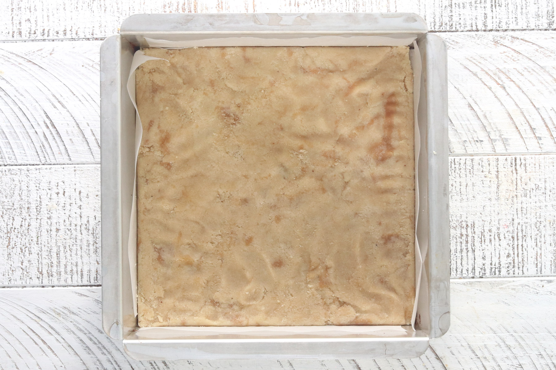 Almond flour base for healthy snickers bars pressed firmly into a parchment-lined square pan, ready for the freezer.