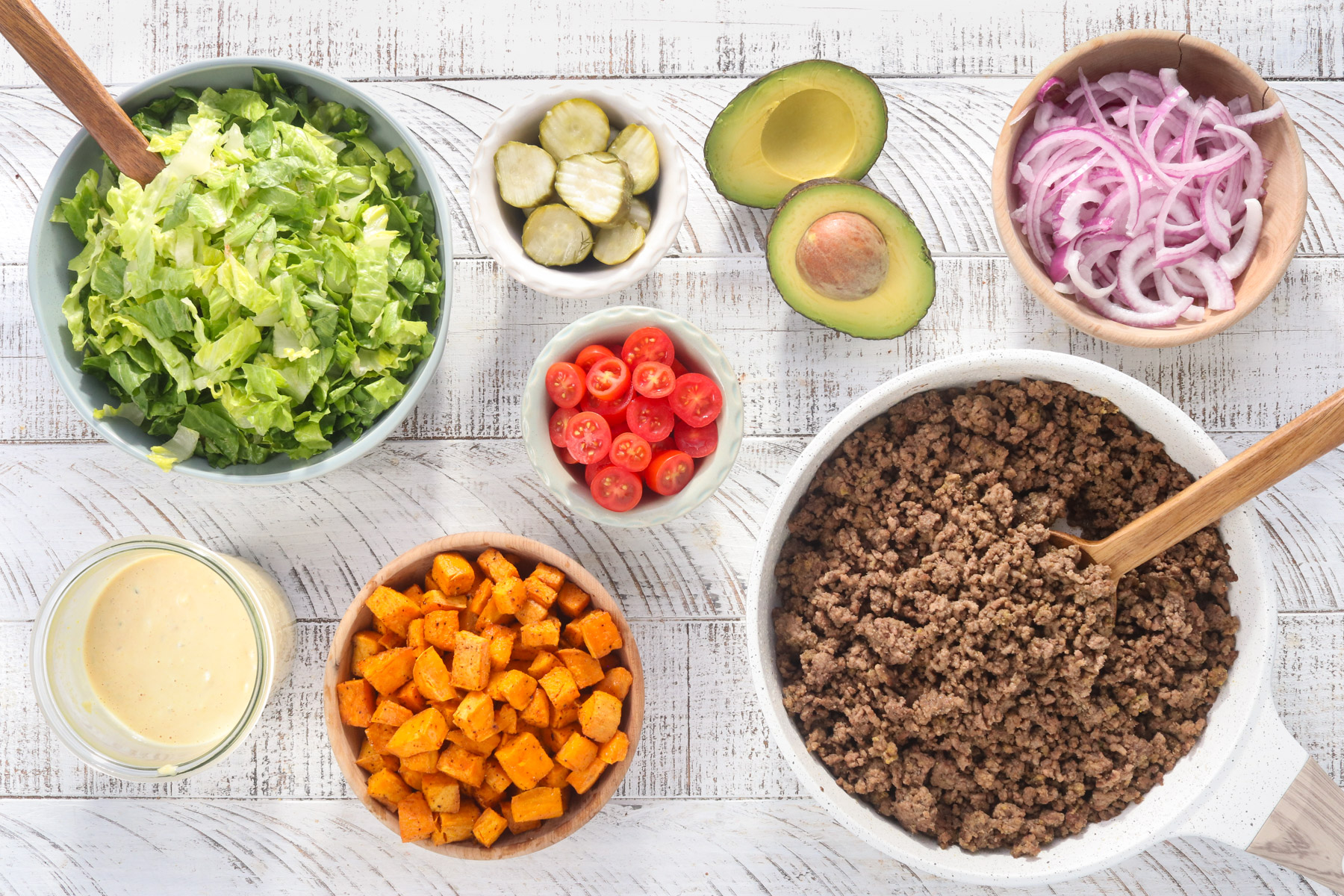 burger bowl ingredients laid out with ground beef, roasted sweet potatoes, lettuce, tomatoes, pickles, red onion, avocado, and burger sauce