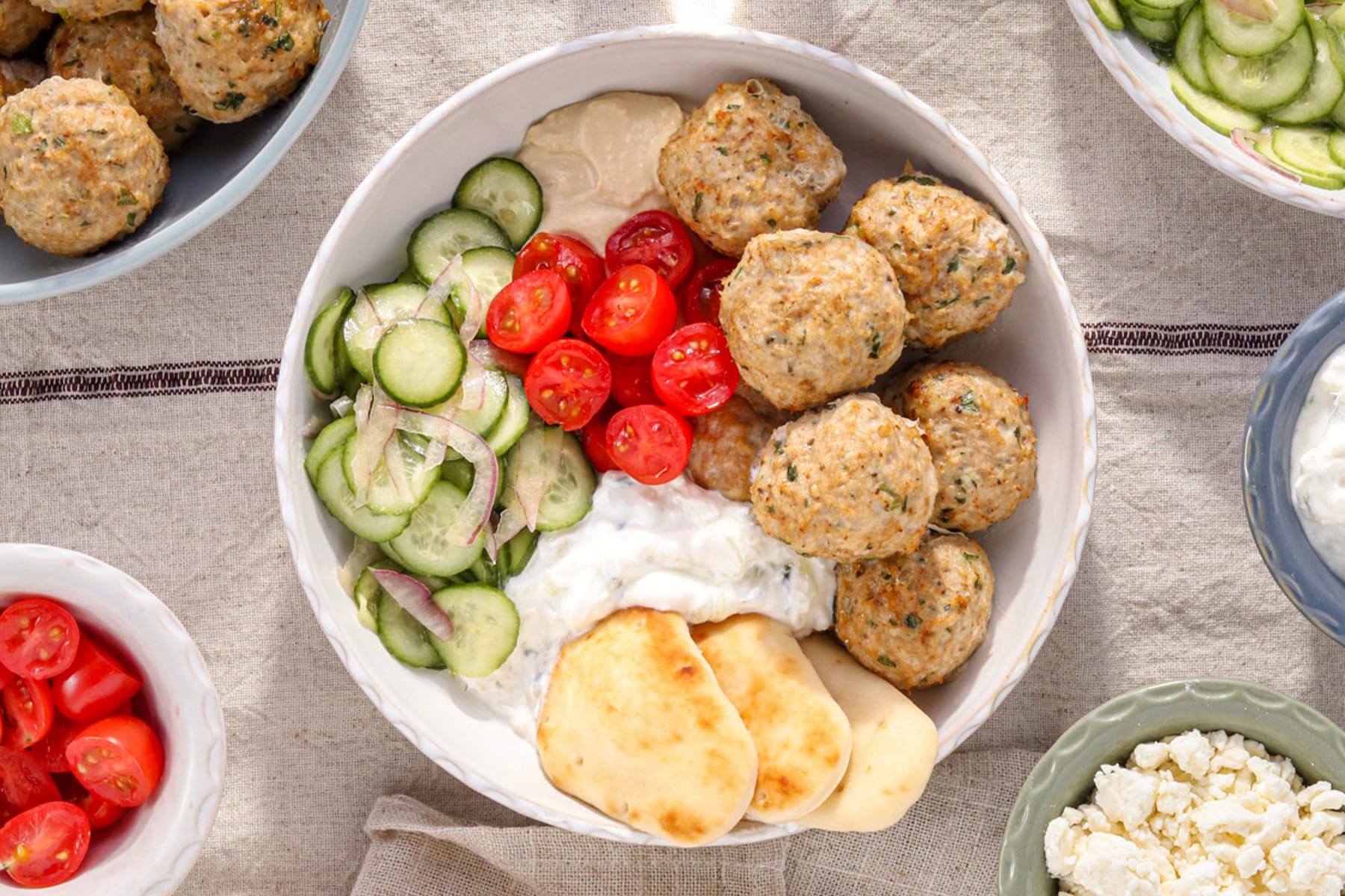 Mediterranean chicken bowl with herby baked chicken meatballs, creamy tzatziki, fresh cucumber salad, cherry tomatoes, hummus, and warm mini naan in a white bowl