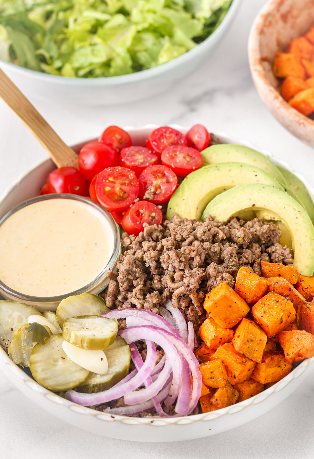burger bowl with ground beef, roasted sweet potatoes, avocado, cherry tomatoes, pickles, red onion, and creamy burger sauce