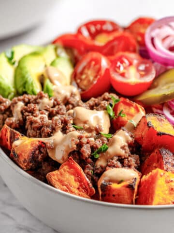 Burger bowl with seasoned ground beef, roasted sweet potatoes, avocado, cherry tomatoes, red onion, pickles, and creamy burger sauce.