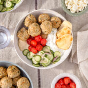 Mediterranean chicken bowl with herby baked chicken meatballs, creamy tzatziki, fresh cucumber salad, cherry tomatoes, hummus, and warm mini naan in a white bowl