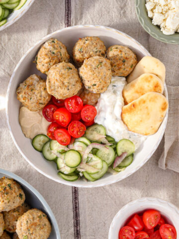 Mediterranean chicken bowl with herby baked chicken meatballs, creamy tzatziki, fresh cucumber salad, cherry tomatoes, hummus, and warm mini naan in a white bowl