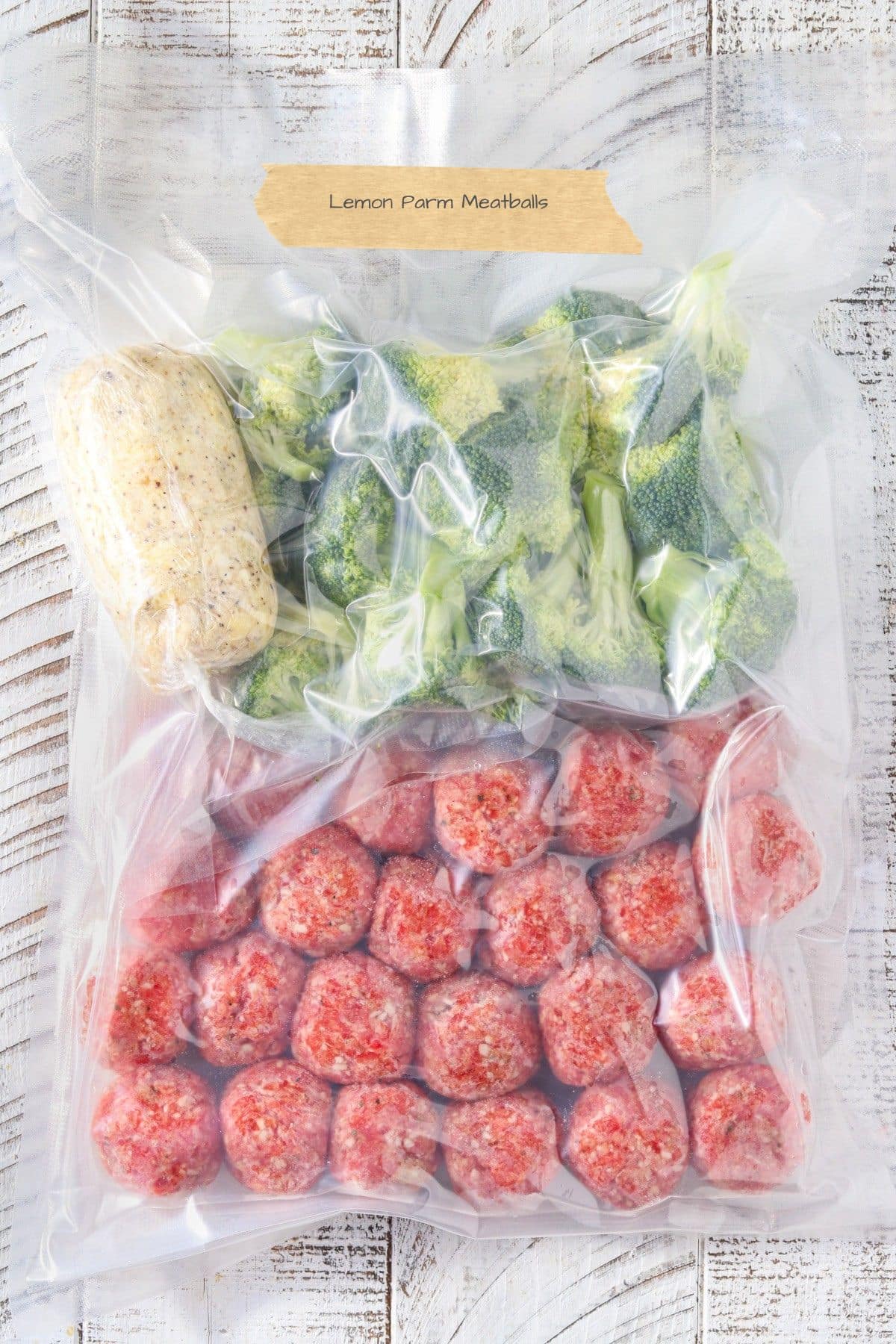Vacuum-sealed freezer meal kit with raw lemon parmesan chicken meatballs, fresh broccoli, and compound butter labeled “Lemon Parm Meatballs” on a white wood background.