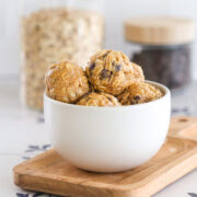 No bake peanut butter power balls with rolled oats and chocolate chips stacked in a white bowl on a wood board, with jars of oats and chocolate chips behind.