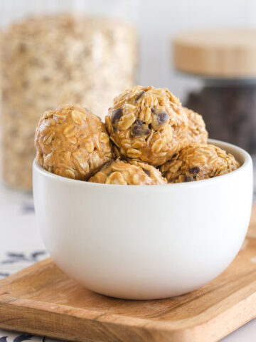 No bake peanut butter power balls with rolled oats and chocolate chips stacked in a white bowl on a wood board, with jars of oats and chocolate chips behind.