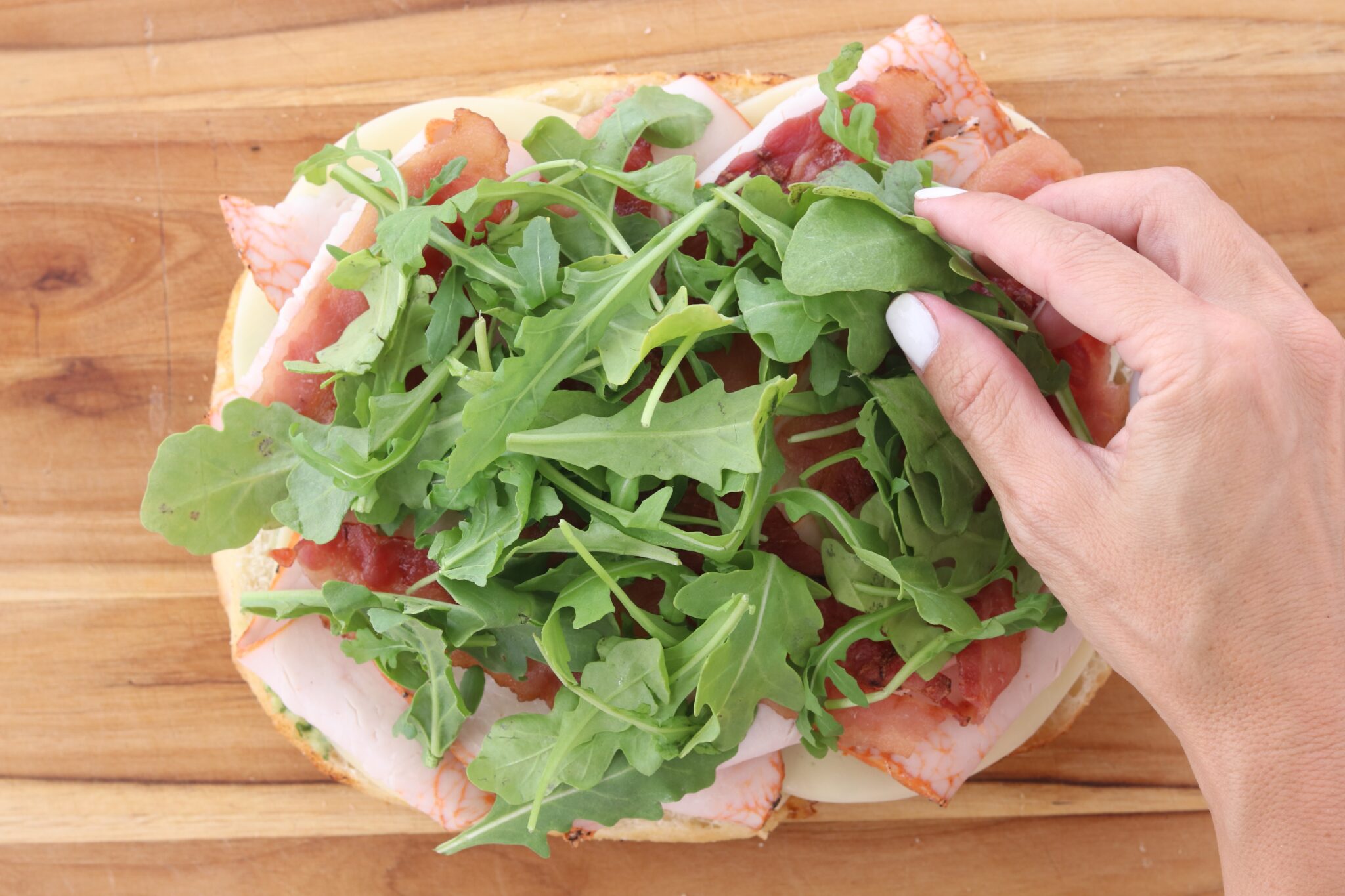 Arugula being added over the turkey, bacon, and provolone on a focaccia sandwich.