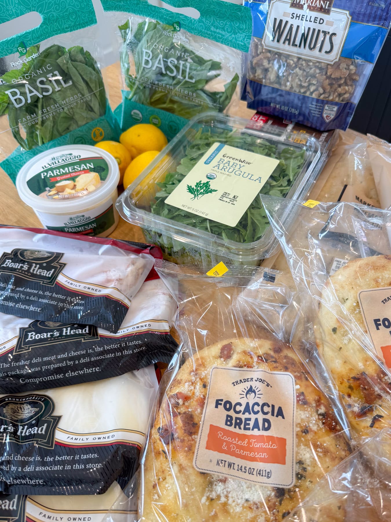 Ingredients for a turkey bacon pesto focaccia sandwich, including focaccia bread, deli turkey, provolone, arugula, basil, parmesan, walnuts, and lemons.