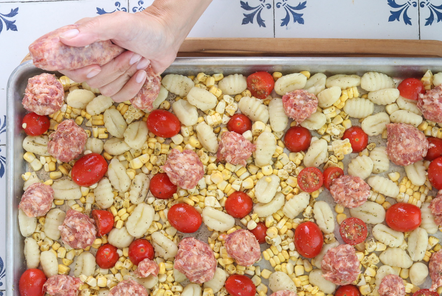 Raw Italian sausage being squeezed from the casing onto a sheet pan with gnocchi, corn and tomatoes before roasting.